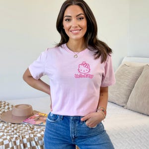 May include: A light pink t-shirt featuring a Hello Kitty graphic in pink. The graphic includes a bow and the text "HelloKitty". The shirt is paired with blue jeans.
