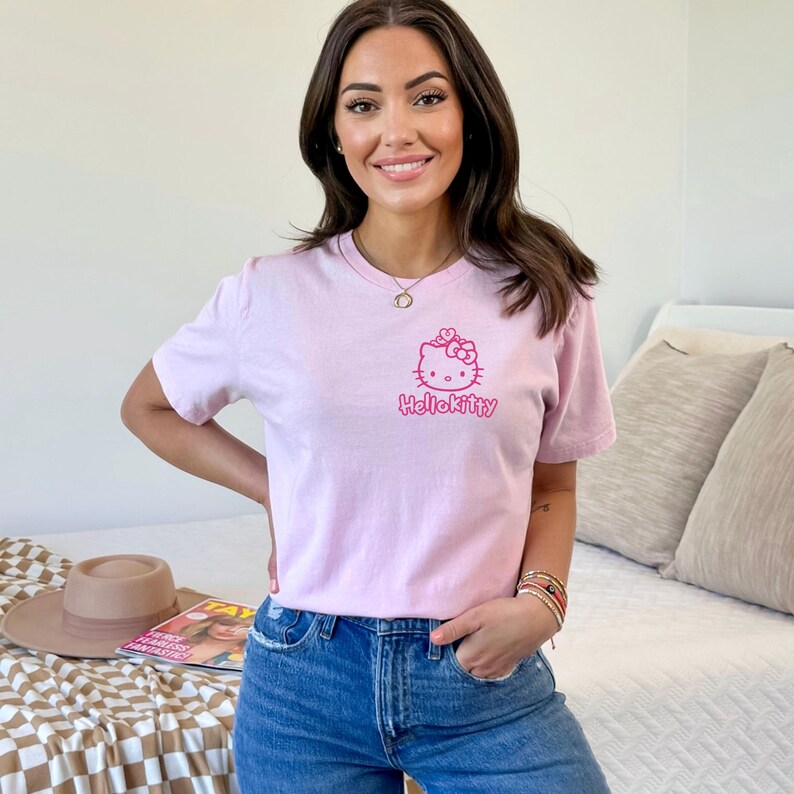May include: A light pink t-shirt featuring a Hello Kitty graphic in pink. The graphic includes a bow and the text "HelloKitty". The shirt is paired with blue jeans.