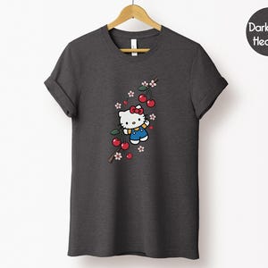 Op de afbeelding: Donkergrijze heather t-shirt met een Hello Kitty-afbeelding. Het ontwerp toont Hello Kitty met een rode strik en blauwe overall, samen met kersen en bloemen. Het t-shirt hangt aan een houten hanger.