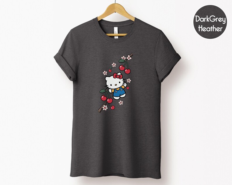 Op de afbeelding: Donkergrijze heather t-shirt met een Hello Kitty-afbeelding. Het ontwerp toont Hello Kitty met een rode strik en blauwe overall, samen met kersen en bloemen. Het t-shirt hangt aan een houten hanger.