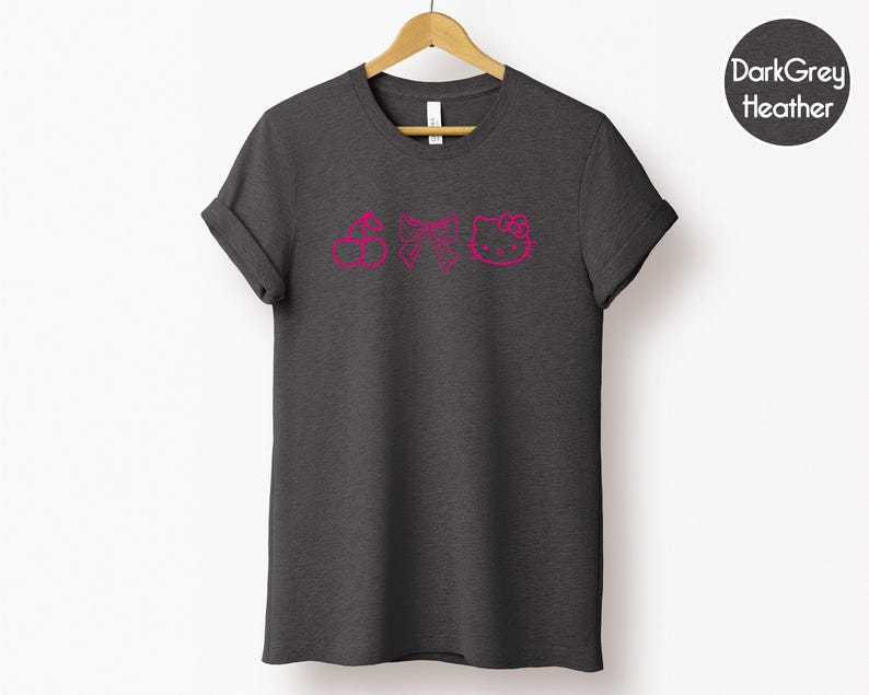 Pu&ograve; includere: T-shirt grigio scuro m&eacute;lange con un design rosa a ciliegia, fiocco e faccina di gatto. La t-shirt &egrave; appesa a una gruccia di legno su sfondo bianco. Le maniche sono arrotolate.