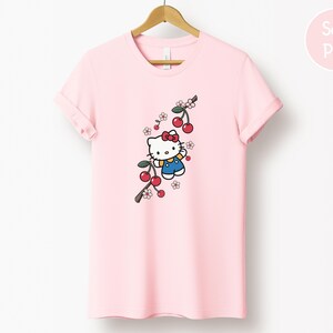 Op de afbeelding: Zachtroze T-shirt met een cartoonfiguur, rode strik, blauwe overall en kersentakontwerp. Het T-shirt hangt aan een houten hanger.