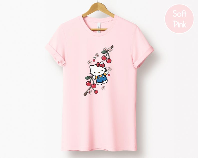 Op de afbeelding: Zachtroze T-shirt met een cartoonfiguur, rode strik, blauwe overall en kersentakontwerp. Het T-shirt hangt aan een houten hanger.
