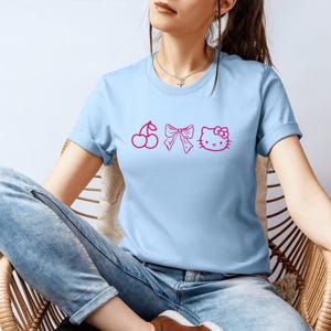 Pu&ograve; includere: T-shirt azzurra con grafica rosa raffigurante una ciliegia, un fiocco e una faccia di gatto. La maglietta ha maniche corte e girocollo. La modella indossa jeans blu.