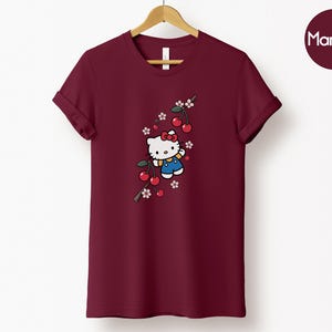 Op de afbeelding: Bordeauxrood T-shirt met een cartoonfiguur, rode strik, blauwe overall en kersenboomtakontwerp. Het T-shirt hangt aan een houten hanger tegen een witte achtergrond.
