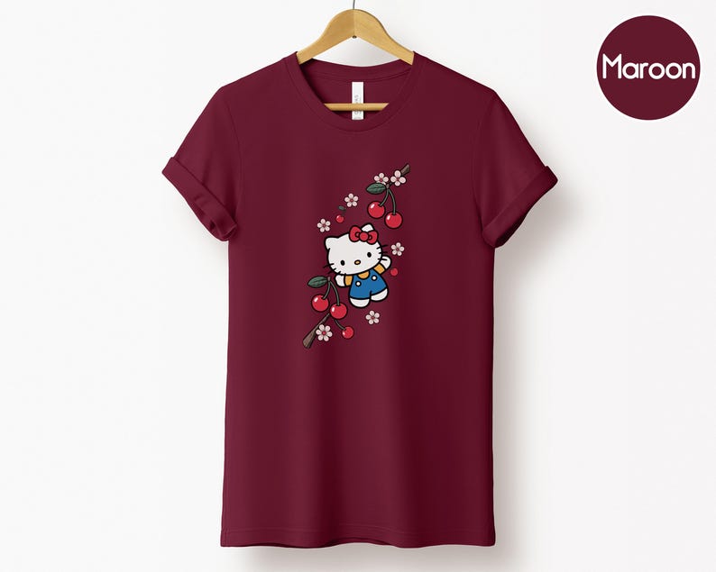 Op de afbeelding: Bordeauxrood T-shirt met een cartoonfiguur, rode strik, blauwe overall en kersenboomtakontwerp. Het T-shirt hangt aan een houten hanger tegen een witte achtergrond.