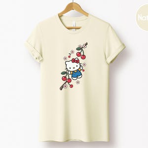 Op de afbeelding: Cr&egrave;mekleurig T-shirt met een stripfiguur met een rode strik en blauwe overall, met een kersenboomtakontwerp. De kersen zijn rood en de bloemen roze en wit. Het shirt hangt aan een houten hanger.