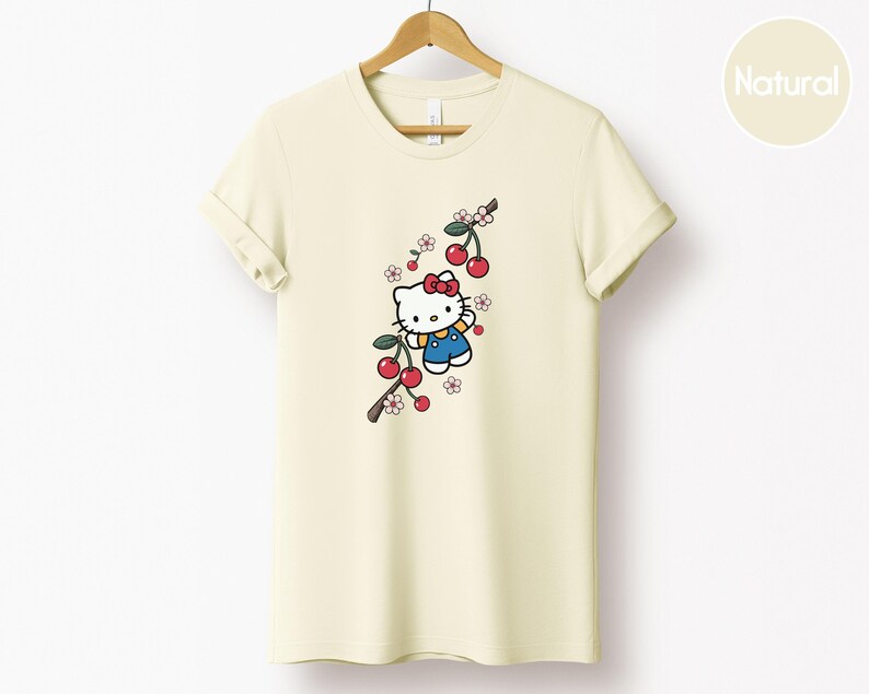 Op de afbeelding: Cr&egrave;mekleurig T-shirt met een stripfiguur met een rode strik en blauwe overall, met een kersenboomtakontwerp. De kersen zijn rood en de bloemen roze en wit. Het shirt hangt aan een houten hanger.