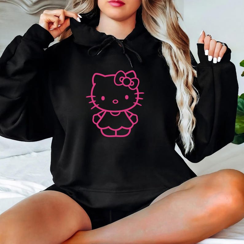 May include: Black hoodie featuring a pink outline of a cartoon cat character with a bow. The hoodie has a drawstring hood and a kangaroo pocket. The cat graphic is centered on the front of the hoodie.