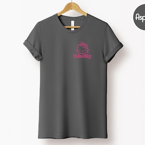 May include: A dark gray t-shirt with rolled-up sleeves. The shirt features a pink Hello Kitty graphic on the chest. The shirt is hanging on a wooden hanger against a white background.