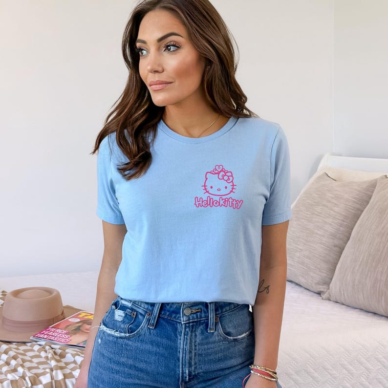 May include: A light blue t-shirt featuring a pink Hello Kitty graphic on the front. The graphic includes the Hello Kitty character and the text "HelloKitty". The shirt is paired with blue denim jeans.