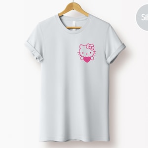 May include: A light gray t-shirt with a pink graphic of a cartoon cat holding a heart. The t-shirt is hanging on a wooden hanger.