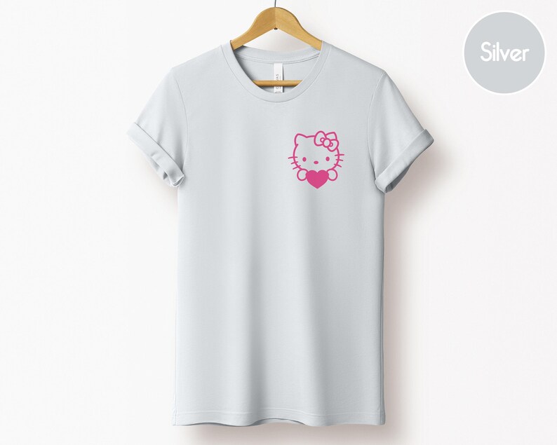 May include: A light gray t-shirt with a pink graphic of a cartoon cat holding a heart. The t-shirt is hanging on a wooden hanger.