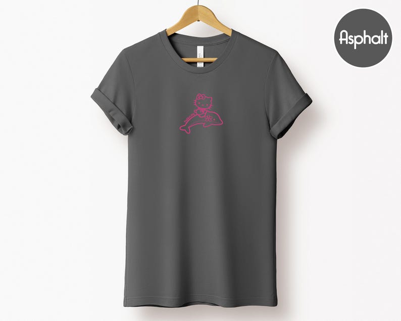 May include: Asphalt-colored t-shirt with a pink graphic of a cartoon character riding a dolphin. The t-shirt is displayed on a wooden hanger against a white background. The sleeves are cuffed. A gray circle with the word "Asphalt" is in the upper right corner.
