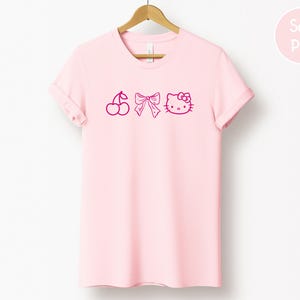 Pu&ograve; includere: Una maglietta rosa chiaro con scollo tondo e maniche arrotolate. La maglietta presenta una grafica magenta di una ciliegia, un fiocco e una faccia di gatto dei cartoni animati. Le parole "Soft Pink" sono in un cerchio rosa in alto a destra.