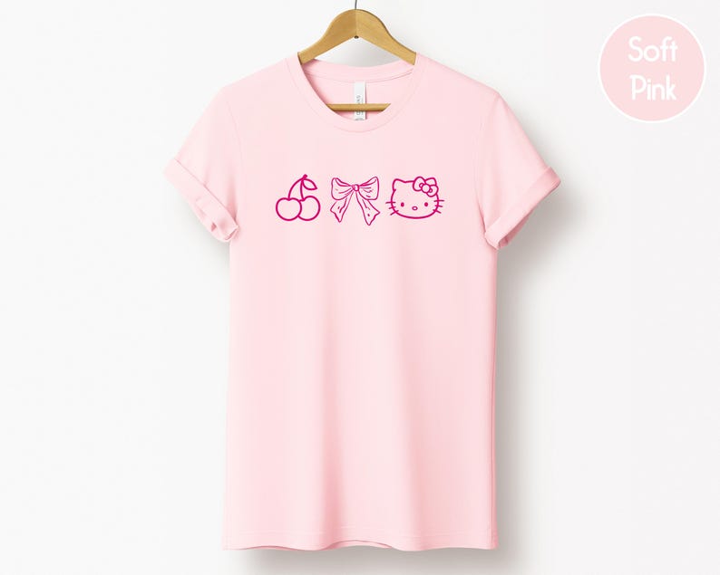 Pu&ograve; includere: Una maglietta rosa chiaro con scollo tondo e maniche arrotolate. La maglietta presenta una grafica magenta di una ciliegia, un fiocco e una faccia di gatto dei cartoni animati. Le parole "Soft Pink" sono in un cerchio rosa in alto a destra.