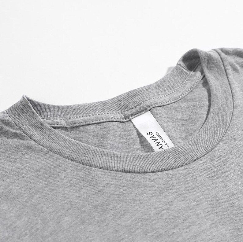May include: Close-up of a heather gray t-shirt neckline. The shirt has a crew neck and a tag that reads "ANVAS LA CANVAS". The fabric appears soft and comfortable, suitable for casual wear.