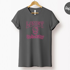 Op de afbeelding: Een donkergrijs t-shirt met opgerolde mouwen. De voorkant toont de roze tekst "LOUEY" boven een Hello Kitty-afbeelding, met "Hellokitty" eronder gedrukt. Het t-shirt hangt aan een houten hanger tegen een witte achtergrond.