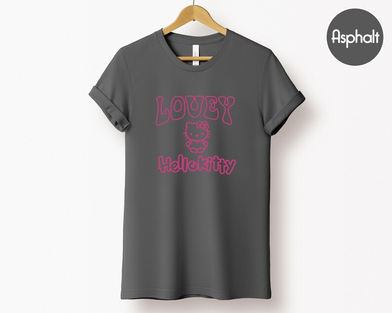 Op de afbeelding: Een donkergrijs t-shirt met opgerolde mouwen. De voorkant toont de roze tekst "LOUEY" boven een Hello Kitty-afbeelding, met "Hellokitty" eronder gedrukt. Het t-shirt hangt aan een houten hanger tegen een witte achtergrond.