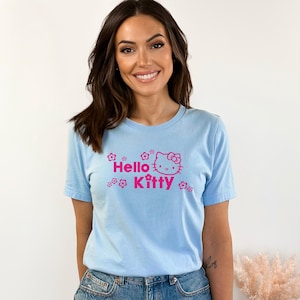 May include: Pale blue t-shirt with the words "Hello Kitty" in pink, accompanied by a Hello Kitty graphic and floral designs. The top has a crew neck.