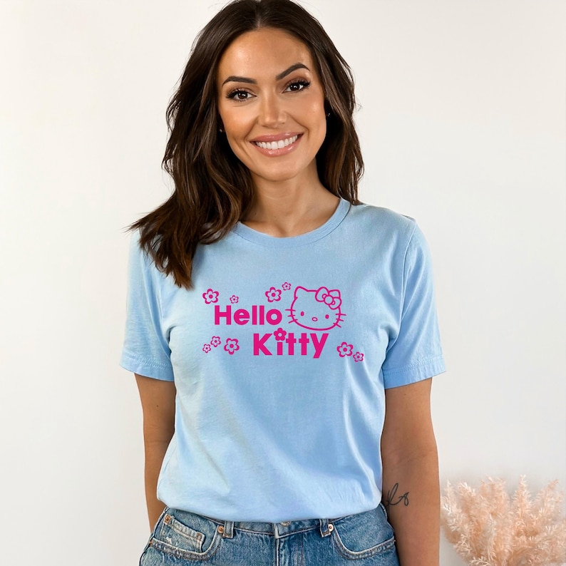 May include: Pale blue t-shirt with the words "Hello Kitty" in pink, accompanied by a Hello Kitty graphic and floral designs. The top has a crew neck.