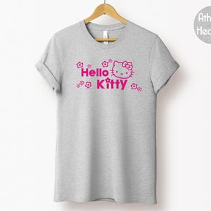 May include: A grey t-shirt with the pink text "Hello Kitty", a graphic of the character's face, and floral designs. The t-shirt is hanging on a wooden hanger.