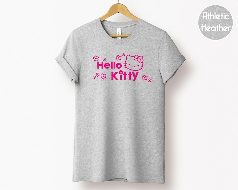 May include: A grey t-shirt with the pink text "Hello Kitty", a graphic of the character's face, and floral designs. The t-shirt is hanging on a wooden hanger.