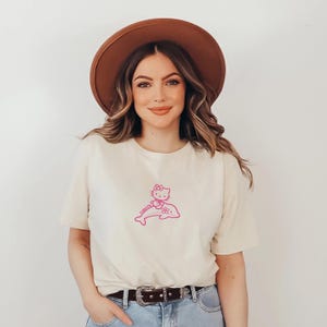 May include: Cream-colored t-shirt featuring a pink graphic of Hello Kitty riding a dolphin. The design is centered on the front of the shirt. The shirt has short sleeves and a crew neck.