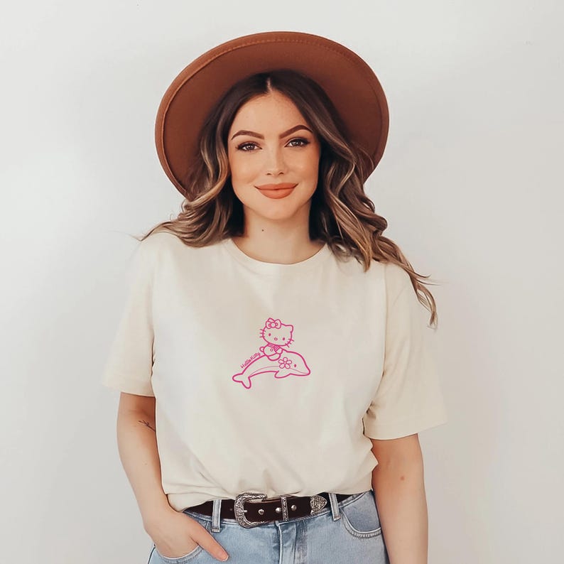 May include: Cream-colored t-shirt featuring a pink graphic of Hello Kitty riding a dolphin. The design is centered on the front of the shirt. The shirt has short sleeves and a crew neck.