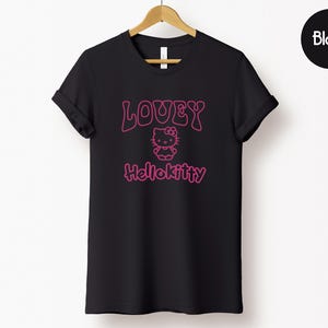 Op de afbeelding: Zwart T-shirt met de tekst "LOVEY HelloKitty" in roze, met een Hello Kitty-afbeelding. Het T-shirt wordt op een houten hanger tegen een witte achtergrond getoond. Het woord "Black" staat in een zwarte cirkel rechtsboven.