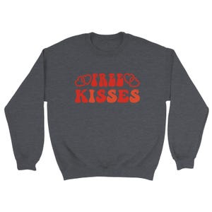 May include: A dark grey sweatshirt featuring the text "FREE KISSES" in a retro font. The words are in shades of orange and red, with heart shapes integrated into the design. A casual, comfortable top.