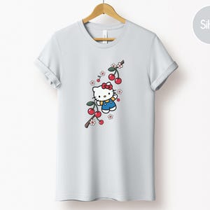 Op de afbeelding: Een wit t-shirt met een cartoonfiguur met een rode strik en blauwe overall, met een kersenboomtakontwerp. De kersen zijn rood en de bloemen roze en wit. Het t-shirt hangt aan een houten hanger.
