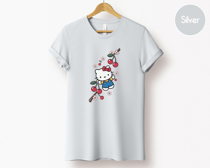 Op de afbeelding: Een wit t-shirt met een cartoonfiguur met een rode strik en blauwe overall, met een kersenboomtakontwerp. De kersen zijn rood en de bloemen roze en wit. Het t-shirt hangt aan een houten hanger.
