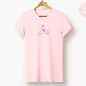 May include: A light pink t-shirt with a rolled sleeve, hanging on a wooden hanger. The shirt features a pink graphic of a cartoon cat riding a dolphin. The words "Soft Pink" are in a pink circle in the upper right corner.