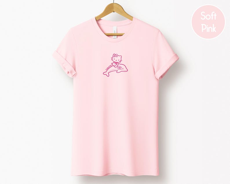 May include: A light pink t-shirt with a rolled sleeve, hanging on a wooden hanger. The shirt features a pink graphic of a cartoon cat riding a dolphin. The words "Soft Pink" are in a pink circle in the upper right corner.