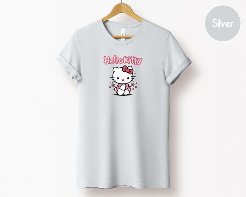 May include: A light gray t-shirt featuring a Hello Kitty graphic. The graphic includes the text "Hello Kitty" in pink above an illustration of the character with a pink bow and flowers.