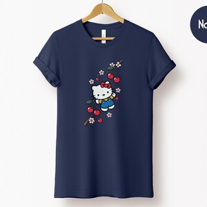 Op de afbeelding: Marineblauw T-shirt met een cartoonfiguur met een rode strik, blauwe tuinbroek en een kersenbloesemtak. De kersen zijn rood en de bloemen roze en wit. Het shirt hangt aan een houten hanger.