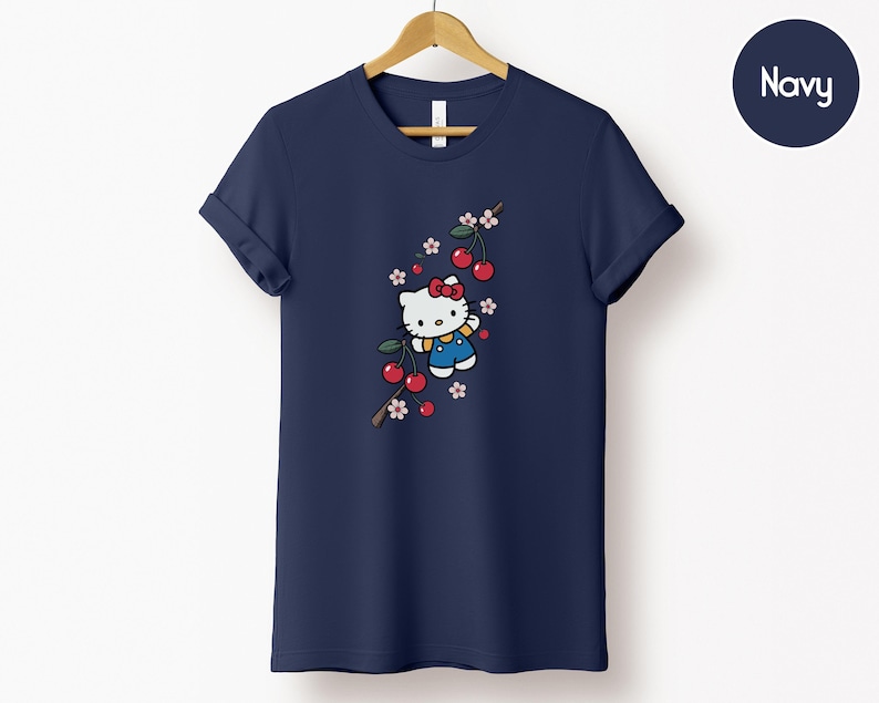Op de afbeelding: Marineblauw T-shirt met een cartoonfiguur met een rode strik, blauwe tuinbroek en een kersenbloesemtak. De kersen zijn rood en de bloemen roze en wit. Het shirt hangt aan een houten hanger.
