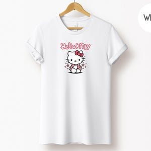 May include: White t-shirt featuring a Hello Kitty graphic. The design includes the text "Hello Kitty" in pink above an illustration of the character with a pink bow and flowers. The shirt is hanging on a wooden hanger.