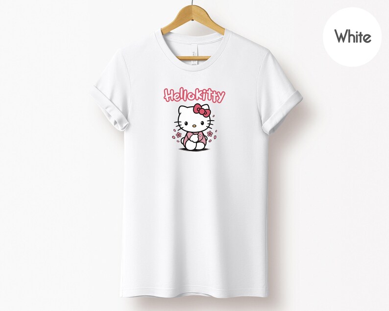 May include: White t-shirt featuring a Hello Kitty graphic. The design includes the text "Hello Kitty" in pink above an illustration of the character with a pink bow and flowers. The shirt is hanging on a wooden hanger.