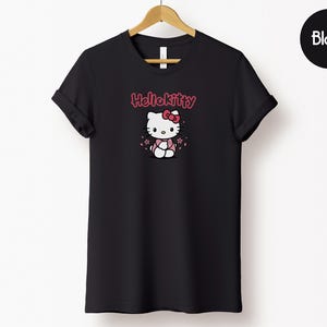 May include: Black t-shirt featuring a Hello Kitty graphic. The design includes the text "Hello Kitty" in pink above an illustration of the character. The shirt is displayed on a wooden hanger against a white background.