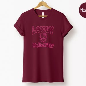 Op de afbeelding: Bordeauxrood T-shirt met opgerolde mouwen, met de tekst "LOUEY" boven een Hello Kitty-afbeelding en de tekst "Hellokitty" eronder. Het shirt hangt aan een houten hanger tegen een witte achtergrond. Een bordeauxrode cirkel met het woord "Maroon" staat rechtsboven.