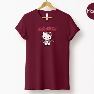 May include: Maroon t-shirt featuring a Hello Kitty graphic. The design includes the text "Hello Kitty" above an illustration of the character. The shirt is displayed on a wooden hanger against a white background.