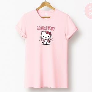 May include: A light pink t-shirt featuring a Hello Kitty graphic. The graphic includes the text "Hello Kitty" above a cartoon image of Hello Kitty with a red bow. The shirt is hanging on a wooden hanger.