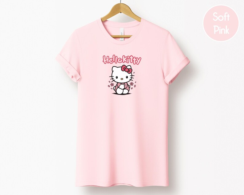 May include: A light pink t-shirt featuring a Hello Kitty graphic. The graphic includes the text "Hello Kitty" above a cartoon image of Hello Kitty with a red bow. The shirt is hanging on a wooden hanger.