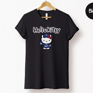 May include: Black t-shirt with a Hello Kitty graphic in a baseball uniform. The text "Hello Kitty" is above the character in white script. The t-shirt is displayed on a wooden hanger against a white backdrop.