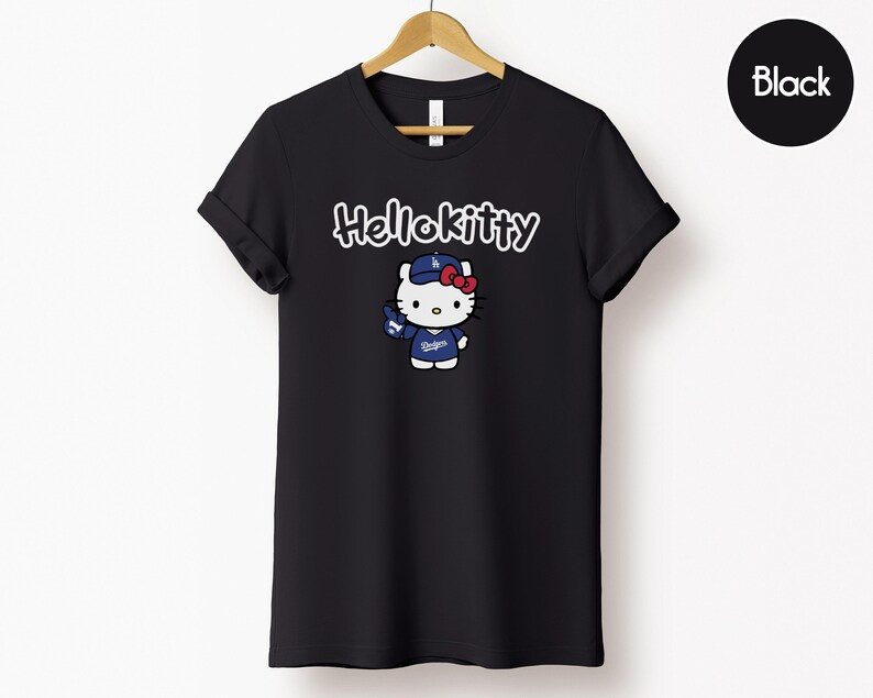 May include: Black t-shirt with a Hello Kitty graphic in a baseball uniform. The text "Hello Kitty" is above the character in white script. The t-shirt is displayed on a wooden hanger against a white backdrop.
