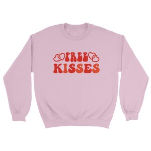May include: A pale pink sweatshirt featuring the text "FREE KISSES" in a retro style. The words are in shades of red and orange, with heart motifs. The sweatshirt has a crew neck and long sleeves.