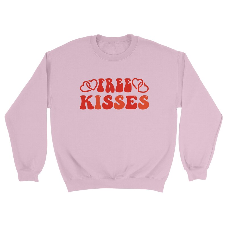 May include: A pale pink sweatshirt featuring the text "FREE KISSES" in a retro style. The words are in shades of red and orange, with heart motifs. The sweatshirt has a crew neck and long sleeves.