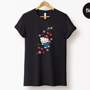 Op de afbeelding: Zwart T-shirt met een cartoonfiguur met een rode strik, blauwe overall en een kersentakontwerp. De kersen zijn rood en de bloemen roze en wit. Het shirt hangt aan een houten hanger.